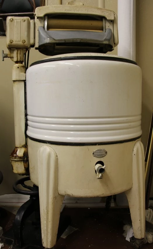 Large white and beige drum with a drain spout in the front bottom. it is supported by 4 metal legs. at the top there appears to be a wringer.