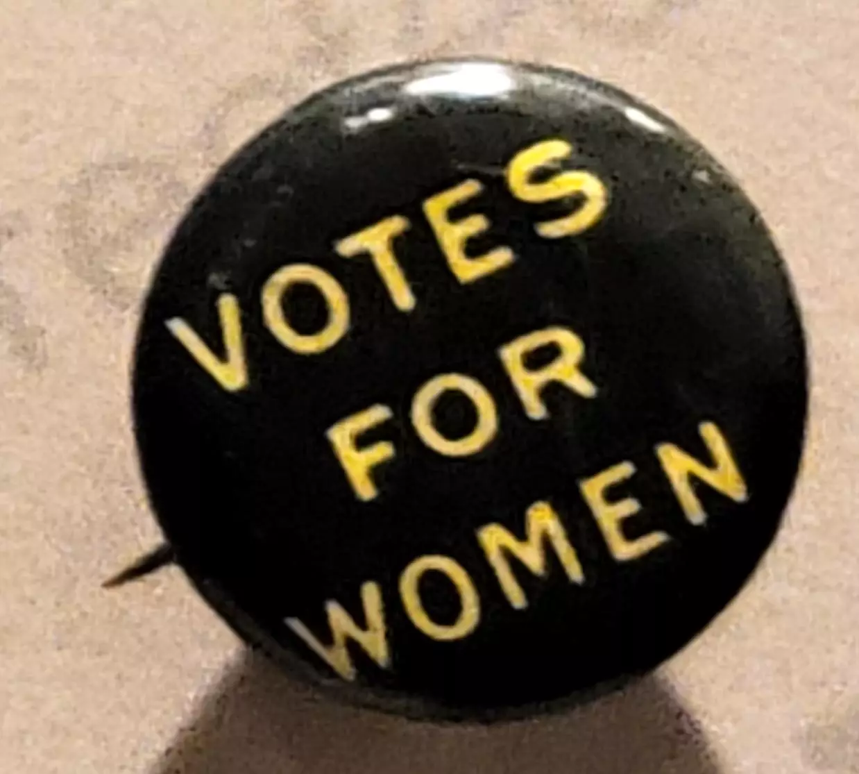 Black metal button with yellow text reading VOTES FOR WOMEN