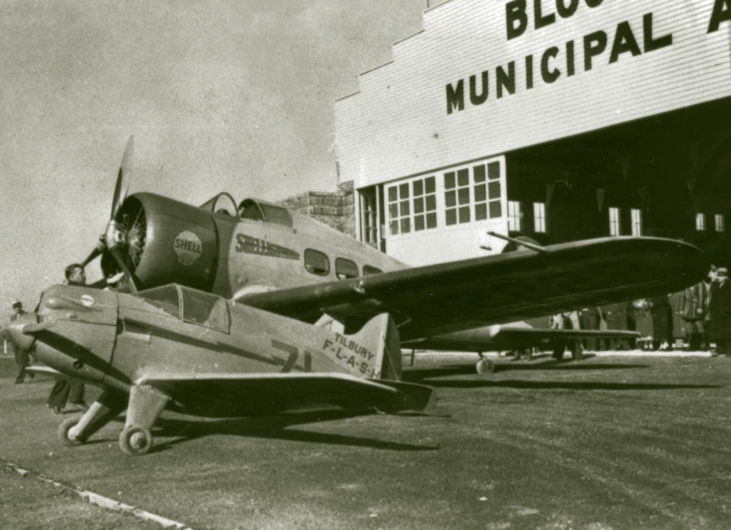 A small airplane is in the foreground. The tail reads tilbury flash behind it is a much larger plane that says shell on the side. they are both parked outside an airplane hangar. words partially cut off but context clues tell us say bloomington municipal airport