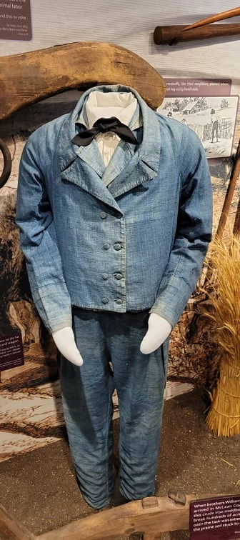 Blue denim jacket with two columns of buttons and a tail, and matching pants on a mannequin. White button-up shirt with a black tie.