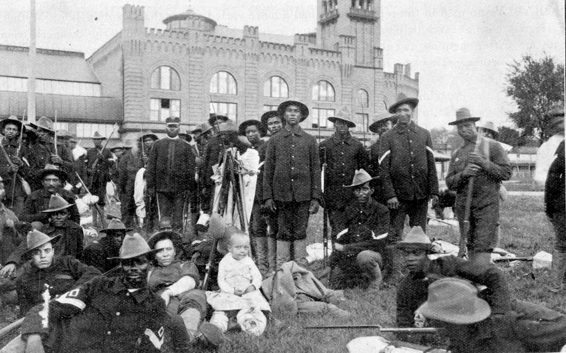 Approximately 30 Black men in hats and uniform, standing, sitting, and laying outside holding guns.