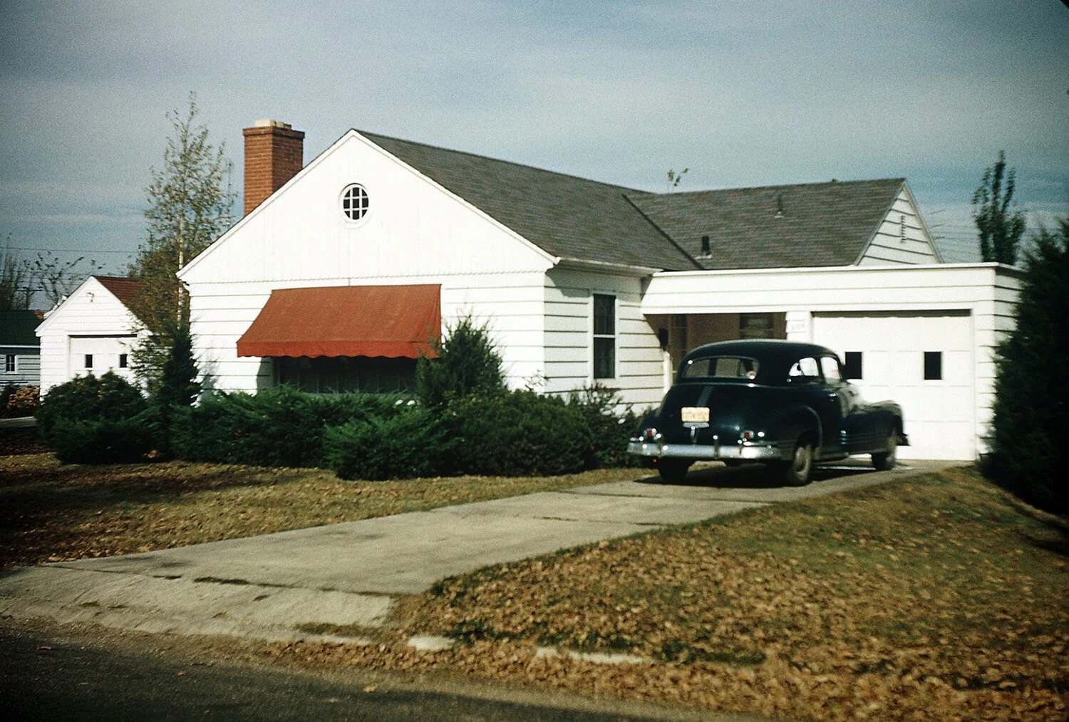 Color photo of a small white house with semi-detached garage and black 1940s-era car in the driveway.