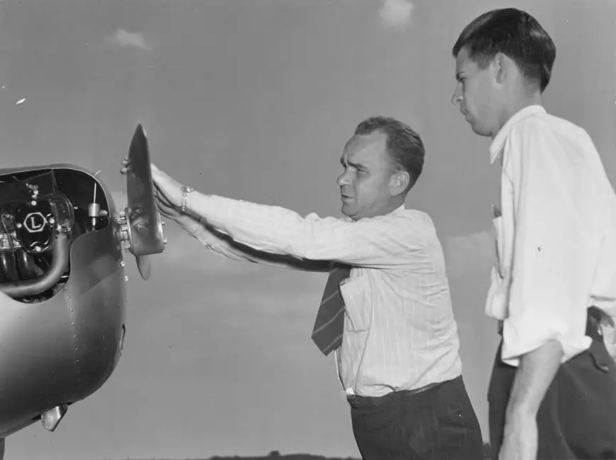 Two light-skinned men wearing white collard shirts are looking at the nose of an airplane, which is eye-level for them. One man has both his hands extended out, touching the top of a propeller blade, presumably about to spin it.