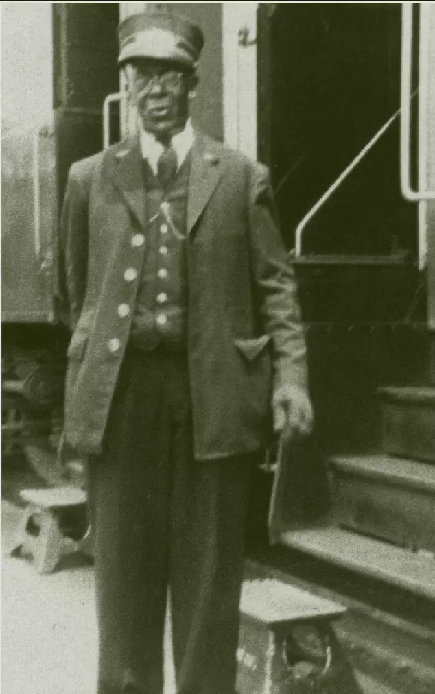An elderly African American Man standing next to a train that is stopped at a station. He is wearing a three piece suit, and the chain of his pocket watch is seen on his vest. He is wearing a porter hat, which is a conical hat with a metal plate on the front, usually with a logo or name of a company, and a small brim.