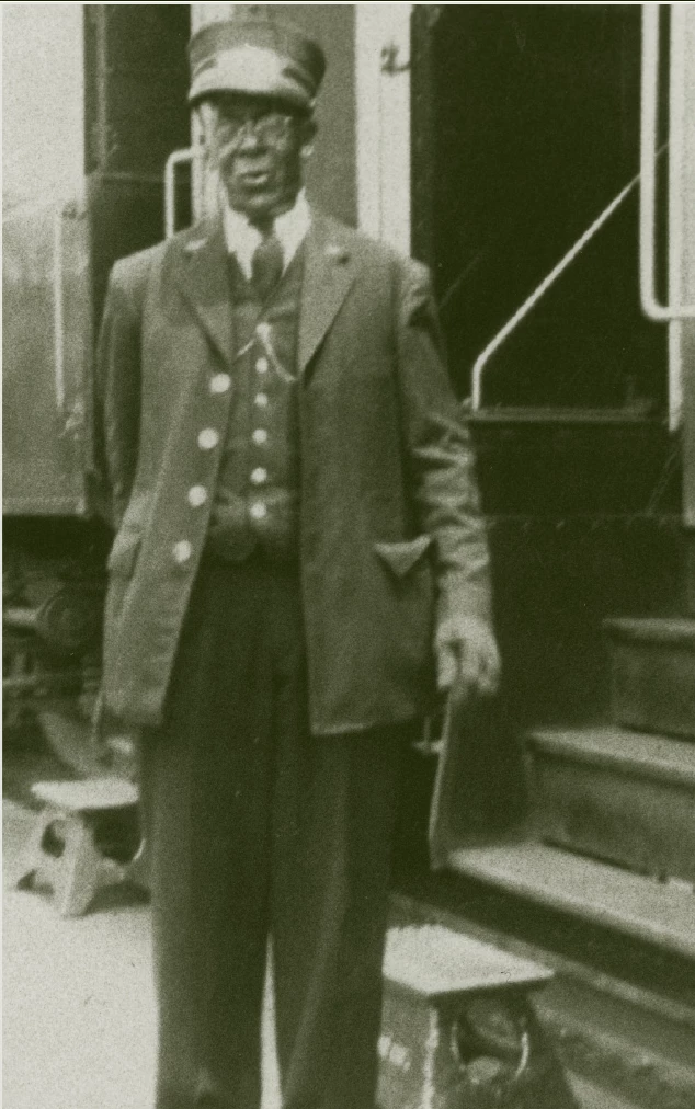An elderly African American Man standing next to a train that is stopped at a station. He is wearing a three piece suit, and the chain of his pocket watch is seen on his vest. He is wearing a porter hat, which is a conical hat with a metal plate on the front, usually with a logo or name of a company, and a small brim.
