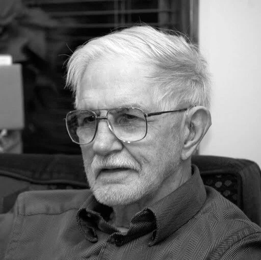 Photograh of an older white man wearing a collared shirt. He is wearing glasses and has a short white beard and white hair.