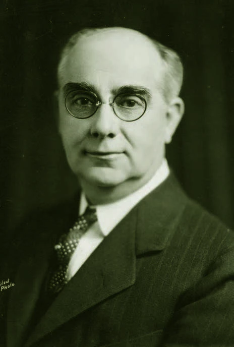 A light-skinned man with balding head and round glasses stares into the camera with a pleasant look. He is wearing a suit and tie.