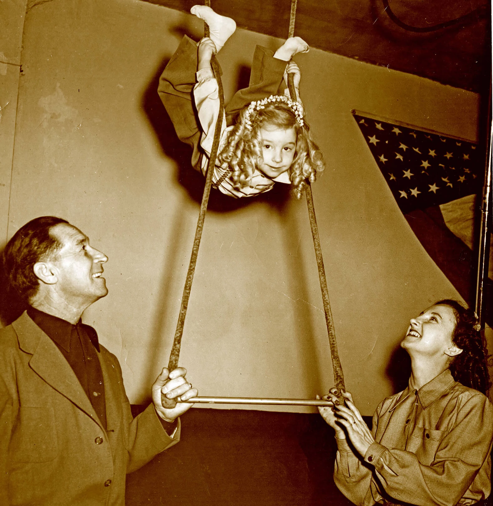 A young girl is holding on two ropes of a trapese, but she is high above the trapese bar. Her feet are above her head and she looks forward with a smile. She has chin-length curly light colored hair and is wearing regular clothes. Her parents are holding the trapese bar, looking up to her, and smiling.
