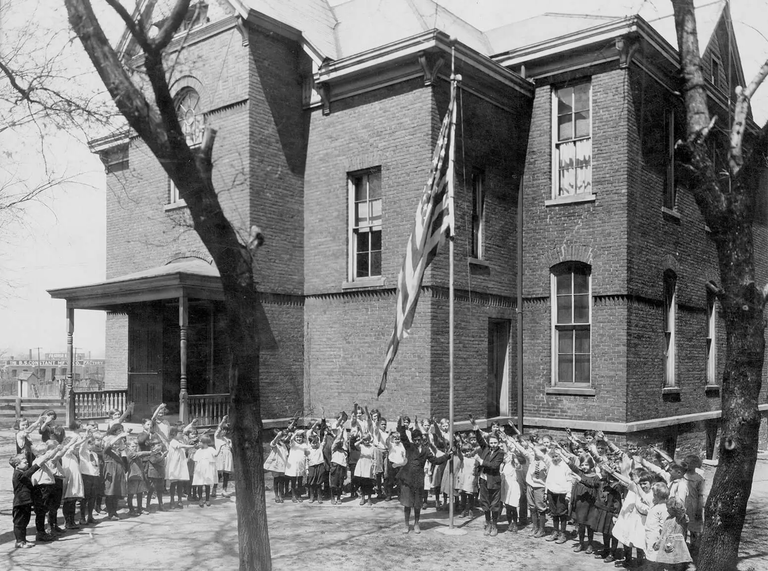 Black and white photo of a large group of children standing outside a two-story brick schoolhouse with a large American flag. They have their hands raised in the air, as if they were presenting the flag for the photo.