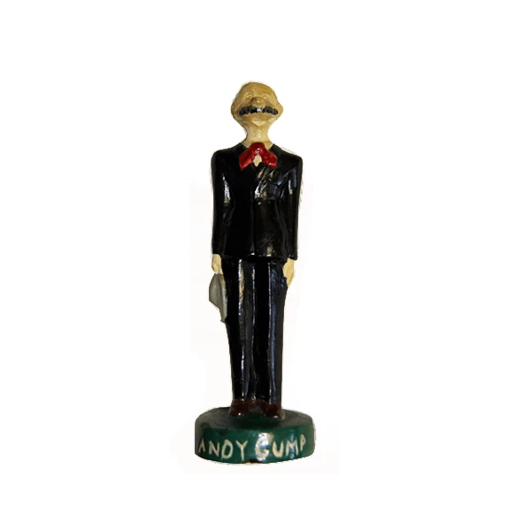A thin light-skinned character with blonde hair, prominent nose, dark mustache, black suit and red bow tie. He is attached to a green base that says