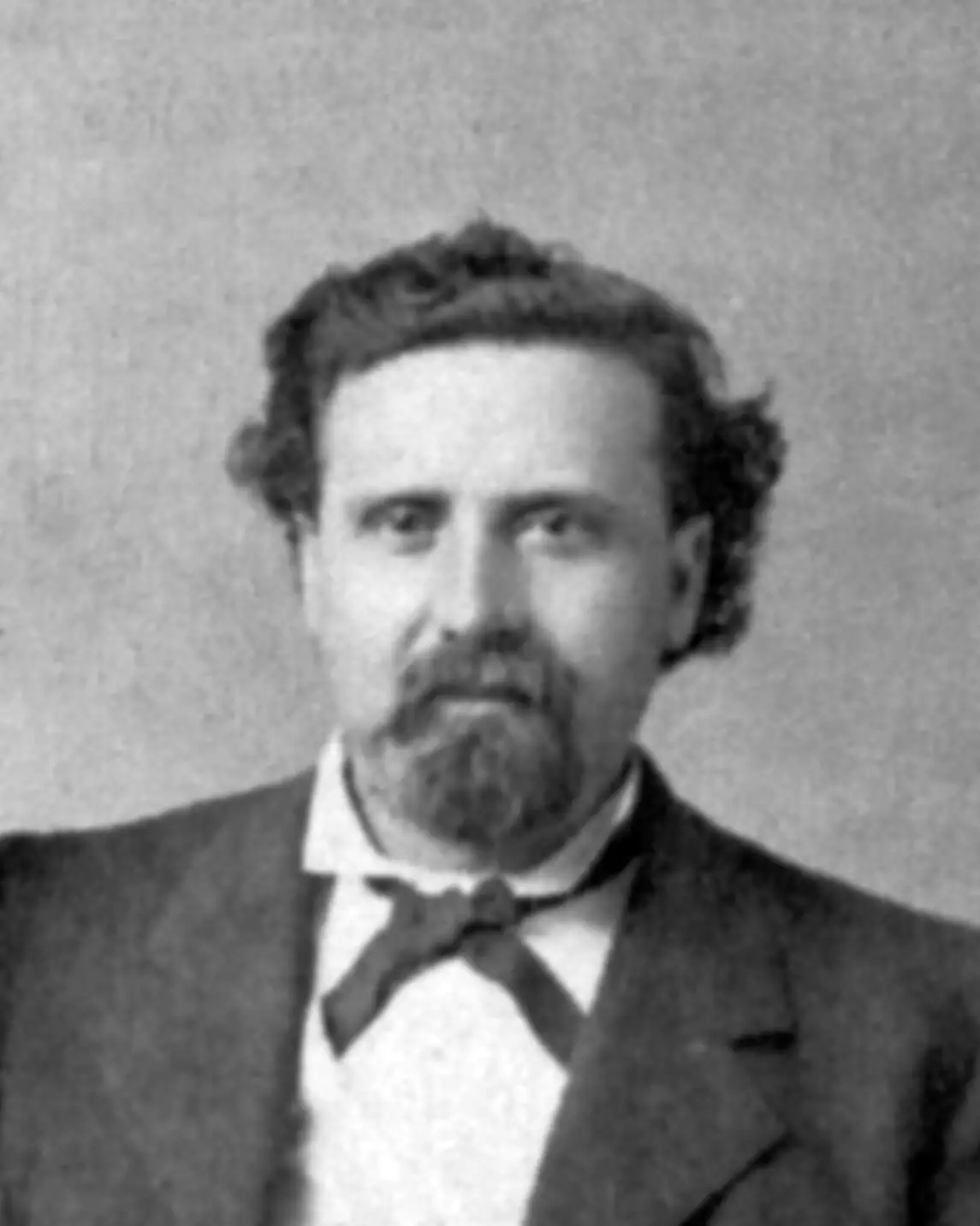 Photo of a light skinned man with dark hair and a goatee. he is wearing a dark suit jacket, white collard shirt, and bow tie.