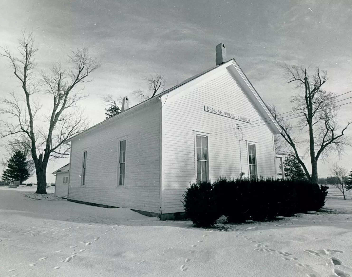 Photo of a small white single-room church surrounded by evergreen bushes and leafless trees in the winter. There is snow on the ground