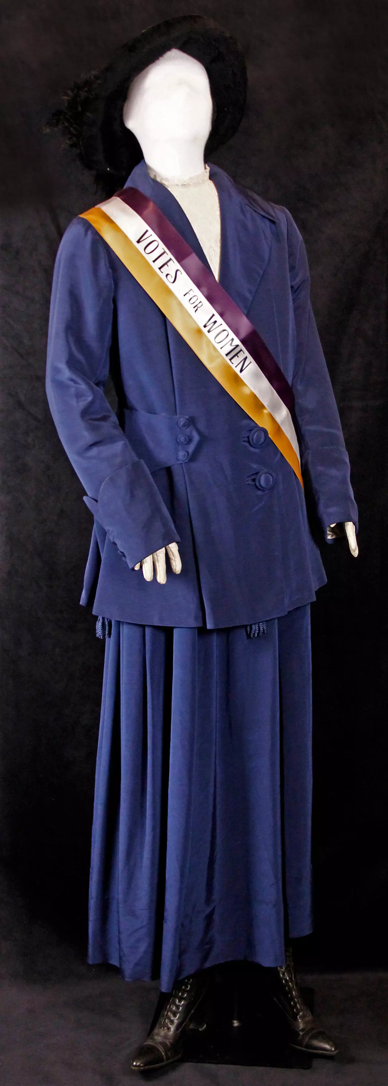 Mannequin dressed in blue velvet women's skirt and jacket, and black hat. A yellow and purple sash reads 'Votes for Women'