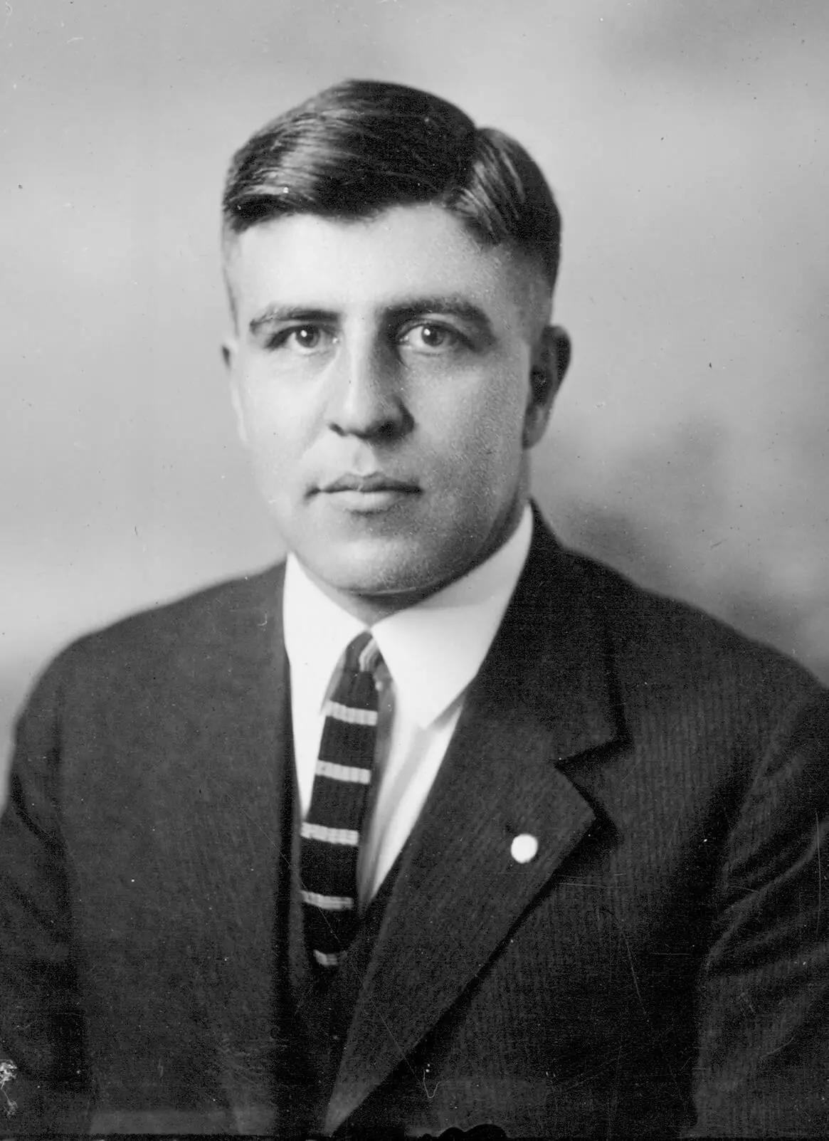 Portrait of a white man with combed dark hair, clean shaven face, striped tie, white shirt and black coat. He is looking into the camera with a straight face.