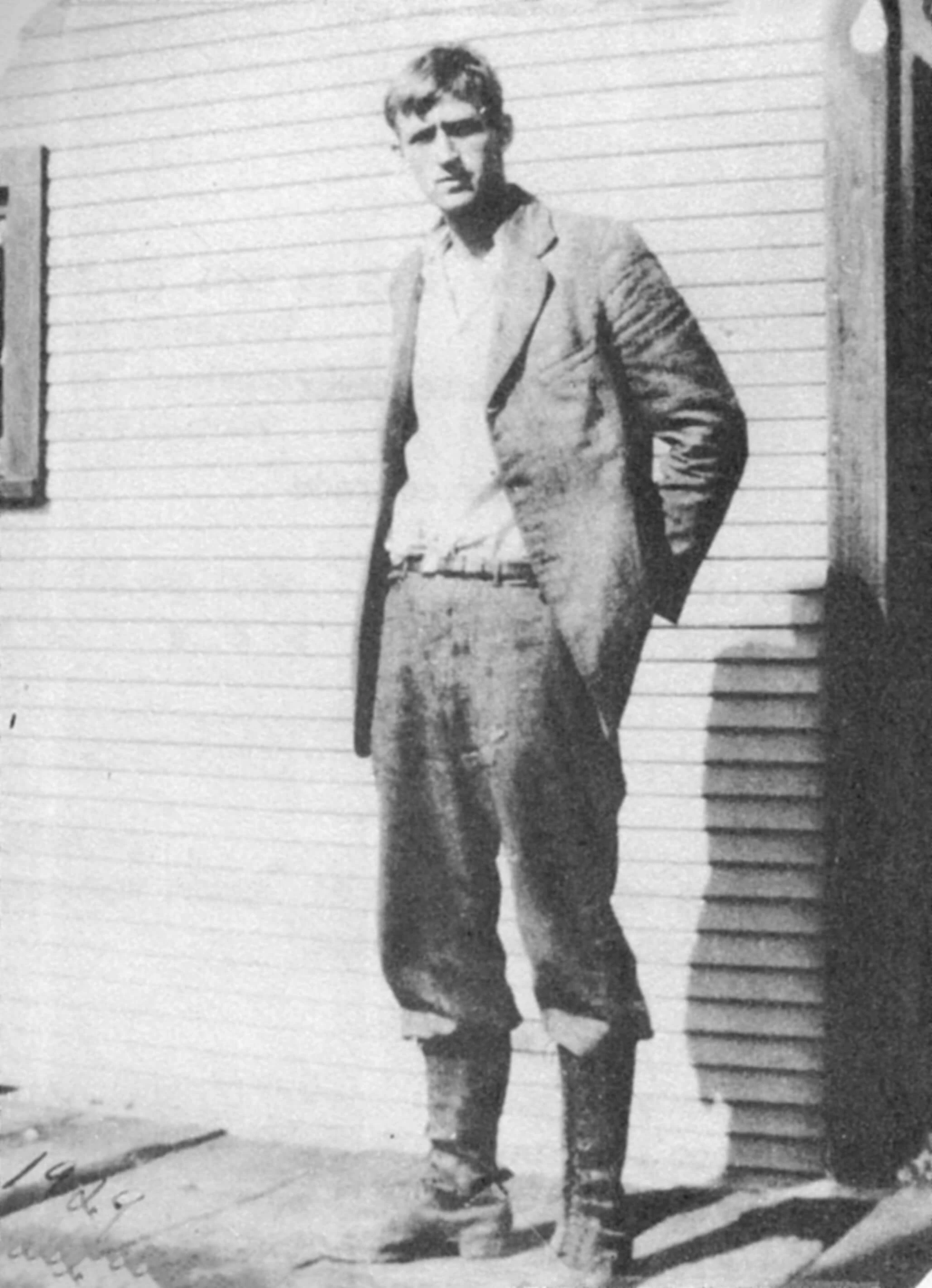 Black and white photo of a young man in a tired looking suit and boots. He is standing, looking at the camera with a straight face, one hand in his back pocket. His clothes look too big.