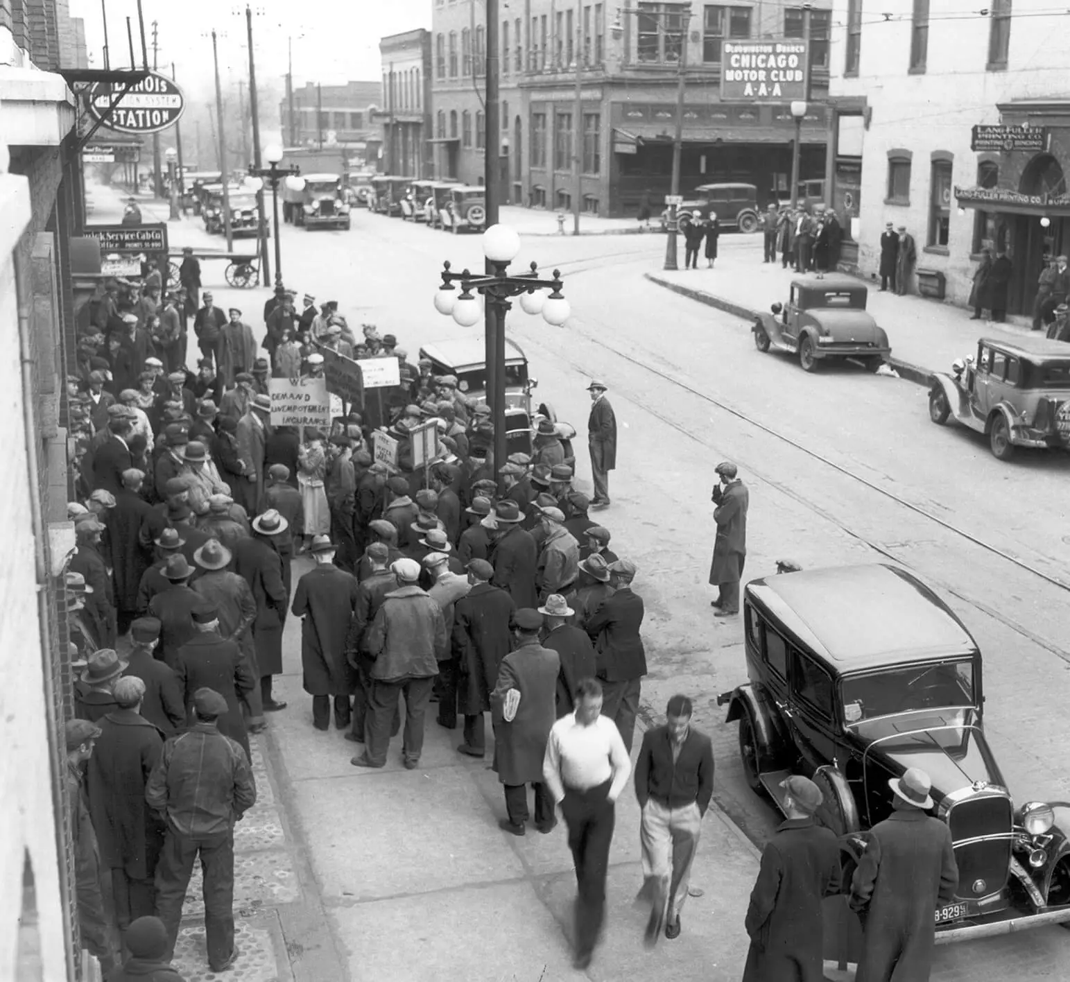 Photograph of a crowd of people on the sidewalk. Almost everyone is wearing hats and long coats. Some appear to be waiting in a line, while others are picketing holding signs.