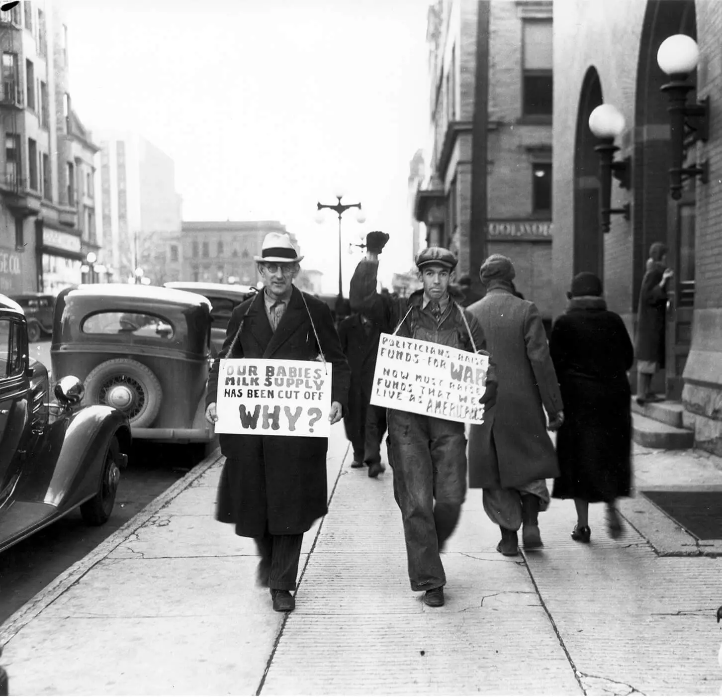 Two white men walk on a sidewalk in Downtown Bloomington with picket signs hanging around their necks. The man on the right is wearing overalls and has his gloved fist raised. Their signs say