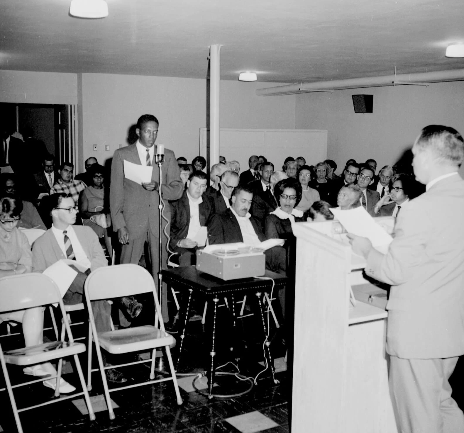 A tall, slim Black man in a suit with a striped tie is speaking at a town meeting. There are dozens of people seated in chairs.