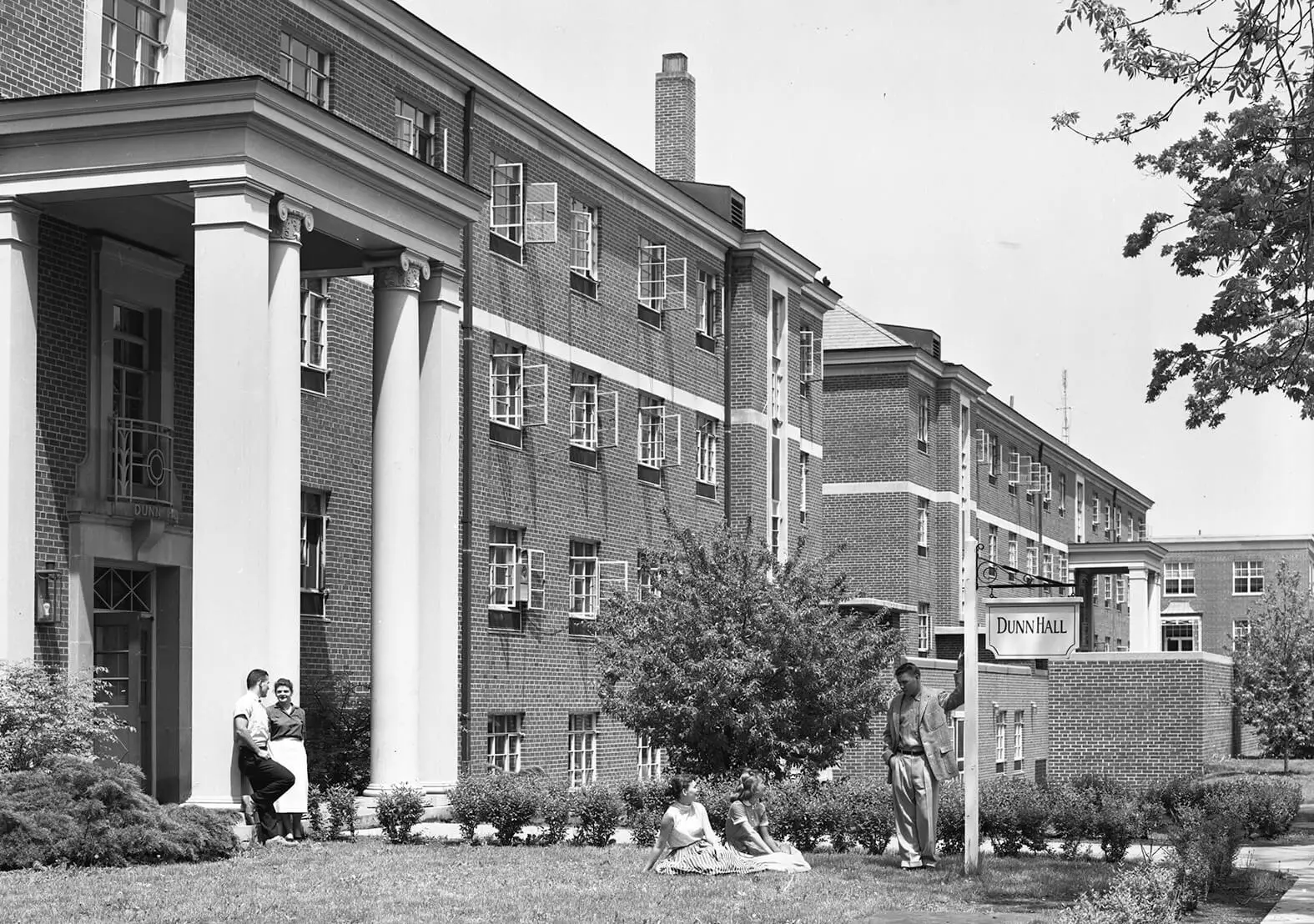 Two brick, four-story residential hall buildings with columns in front. A group of young men and women are outside.