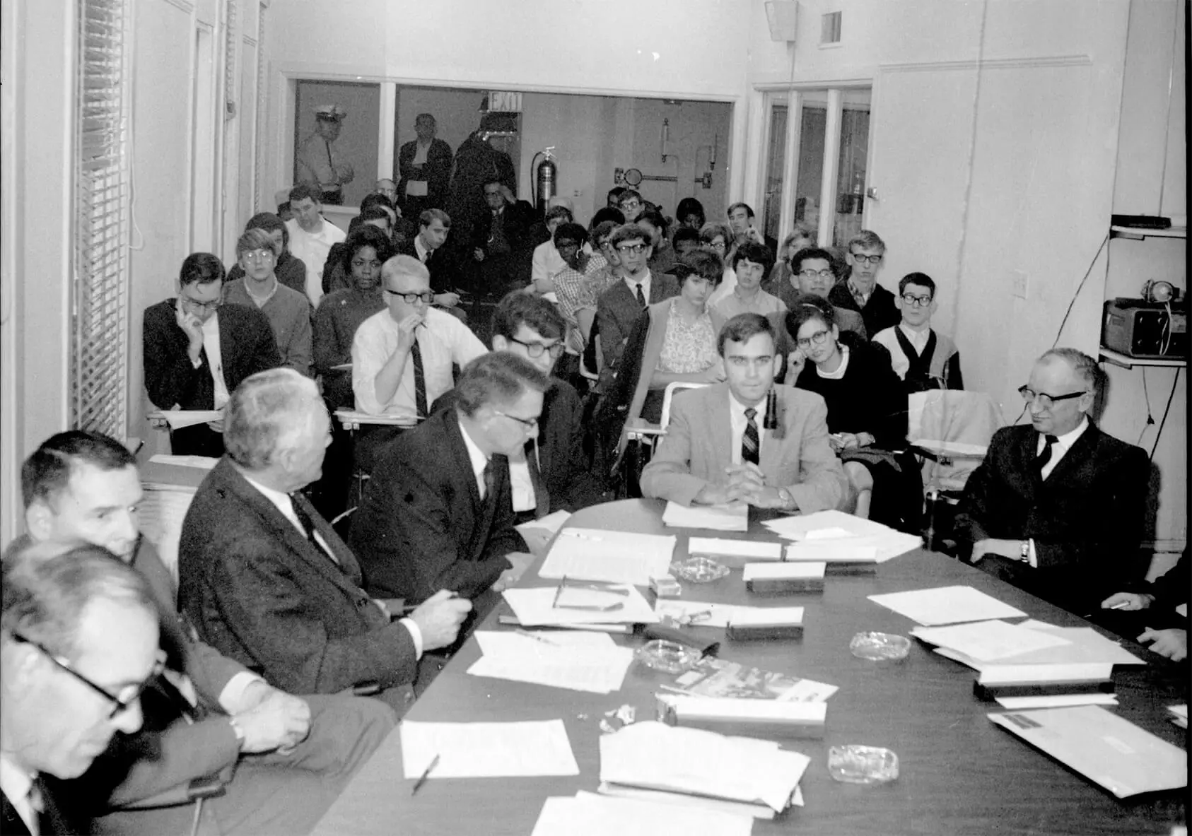 A group of older white men dressed in suits, seated at a conference table with papers scattered around. In the background are 20 or so seated individuals.
