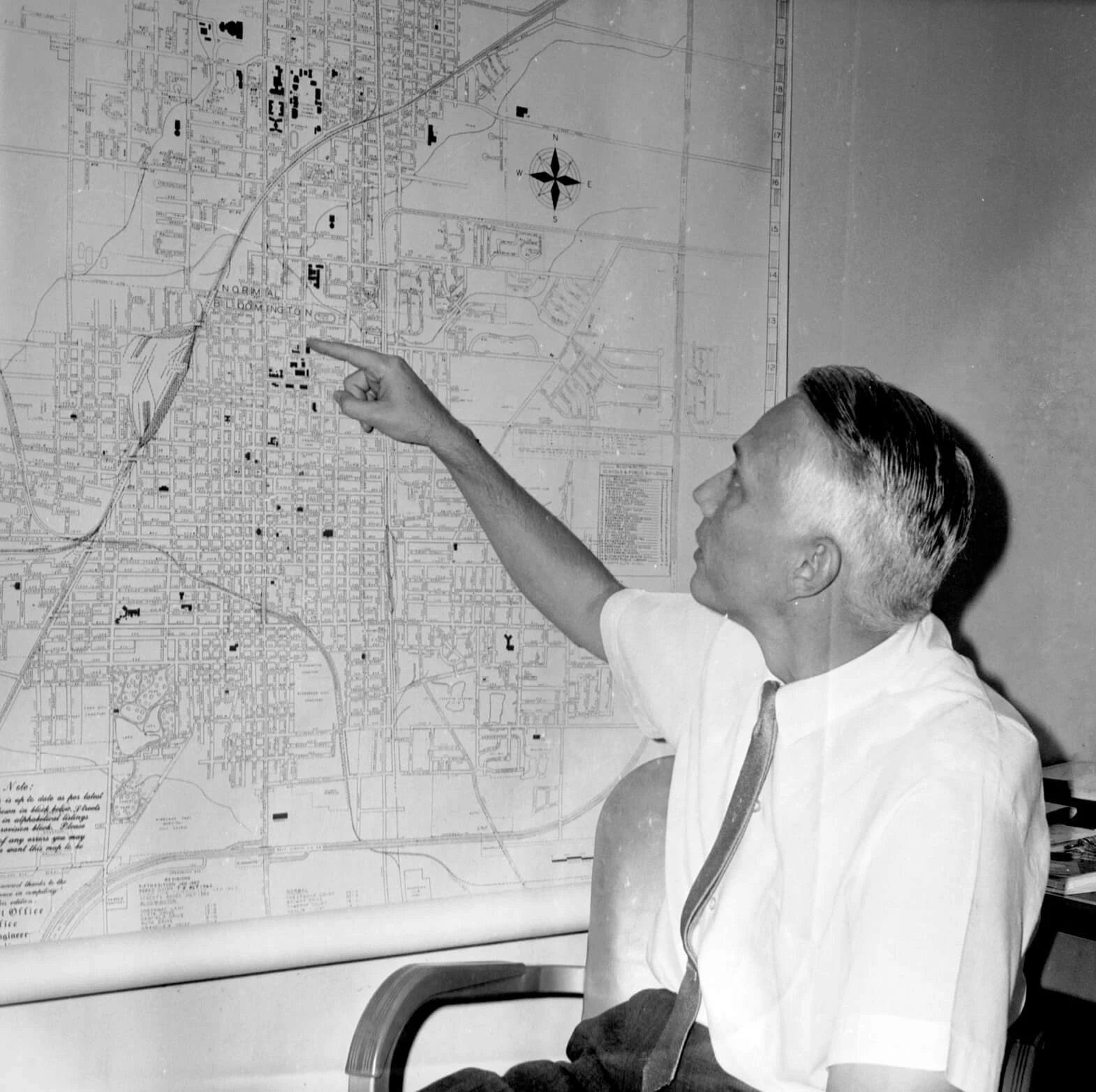 A white man wearing a short sleeved white shirt and tie points to a big map on a wall, showing welcoming neighborhoods on a map of Bloomington-Normal.