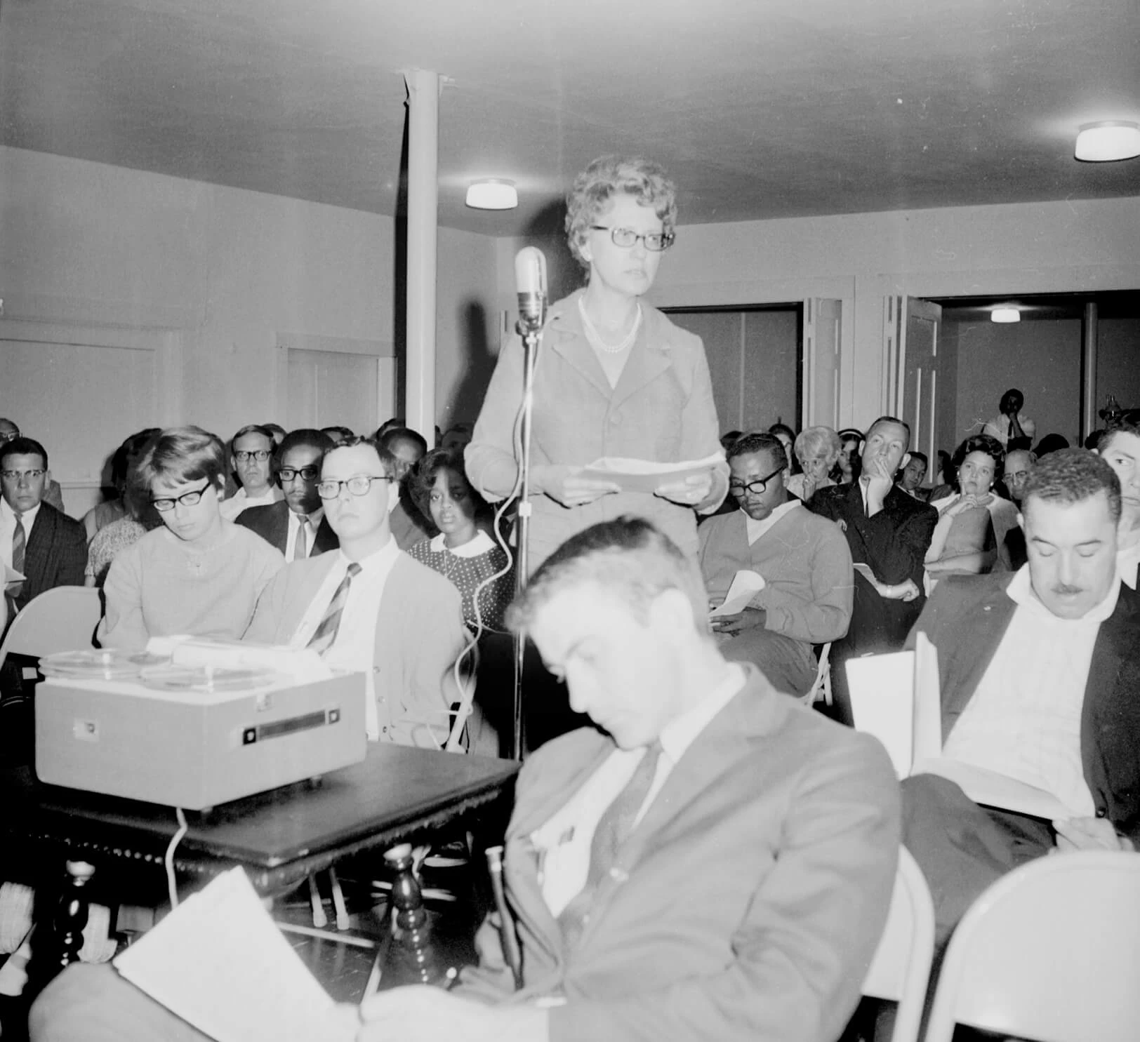 A middle-aged white woman with short curled hair is holding a stack of paper near a microphone at a town meeting. Dozens of people are seated around her.