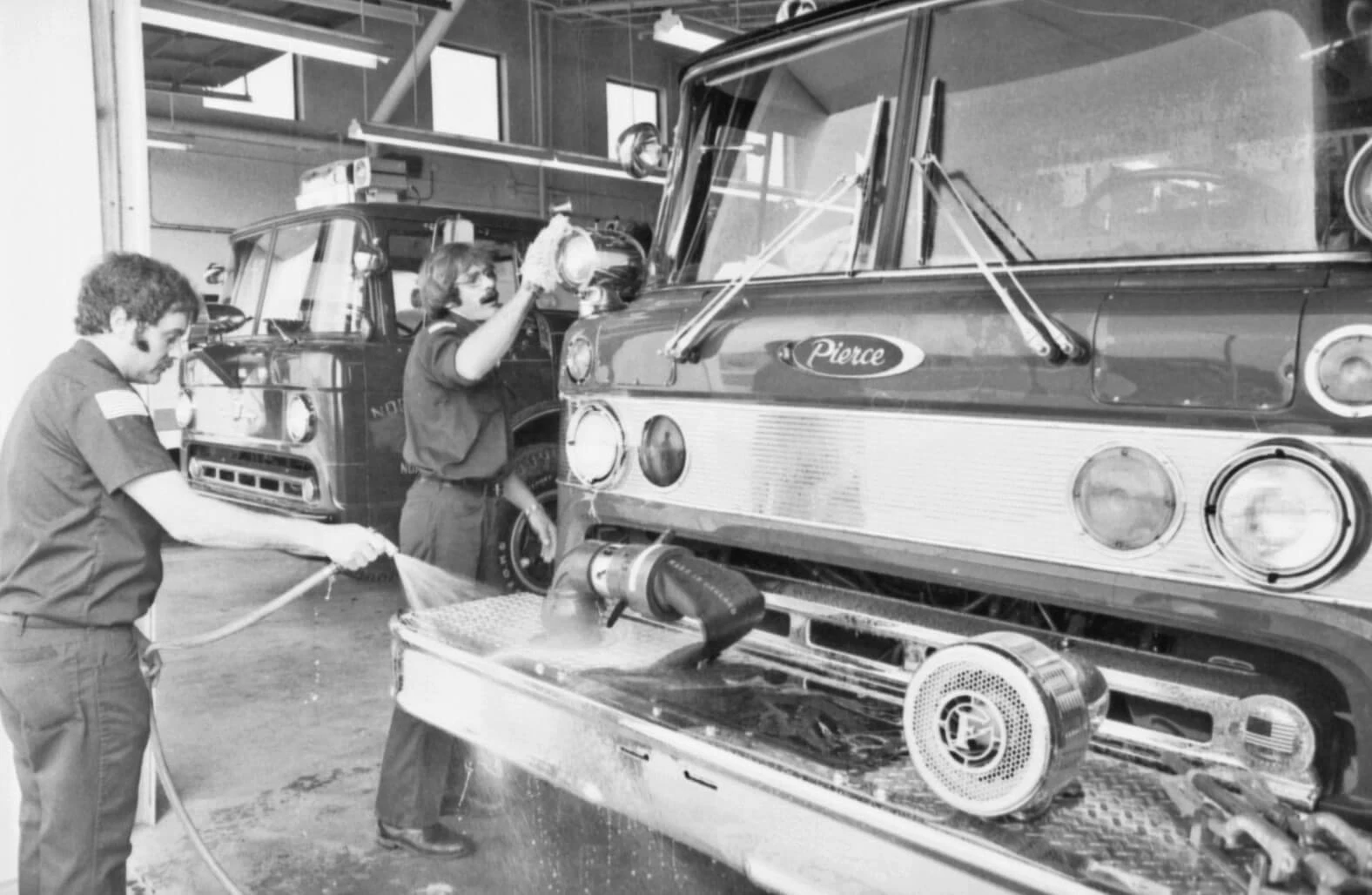 B&W photo of two white men with mustaches washing and polishing a fire engine inside the station.