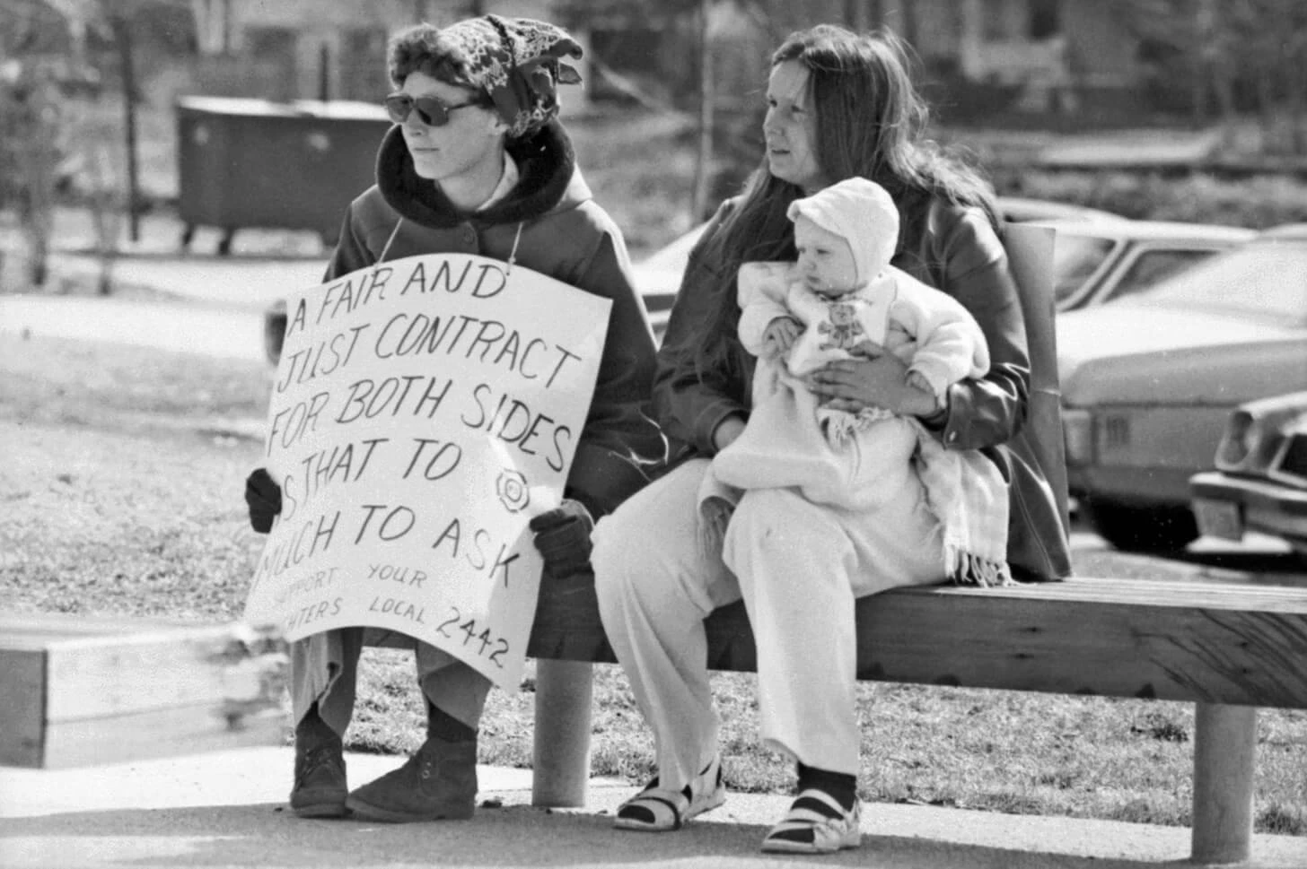 A woman with a posterboard sign around her neck and another woman holding a baby sit on a bench.