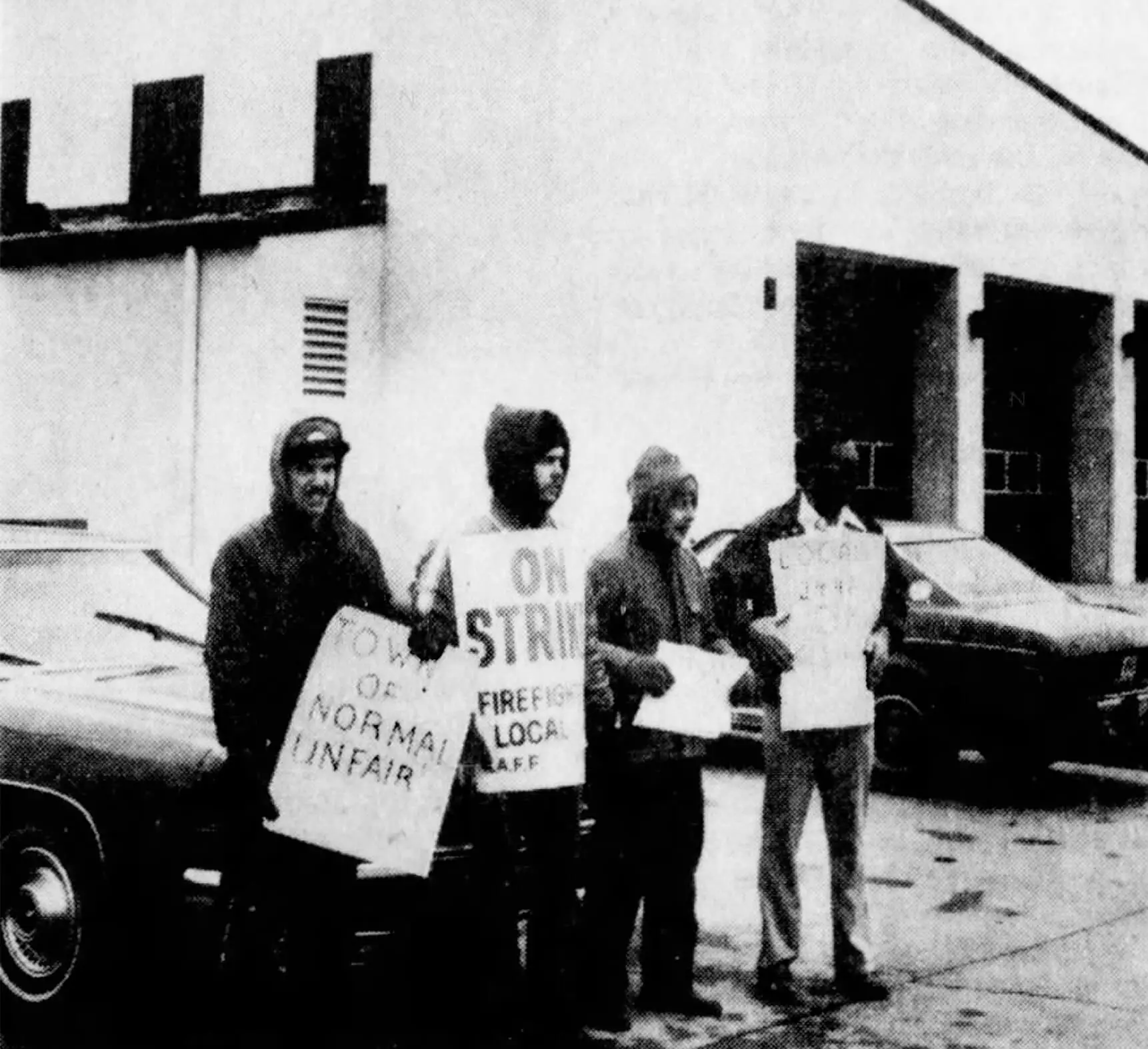 Black and white photo of four men in jackets standing in front of parked cars outside of a fire station while holding posterboard signs. Signs read