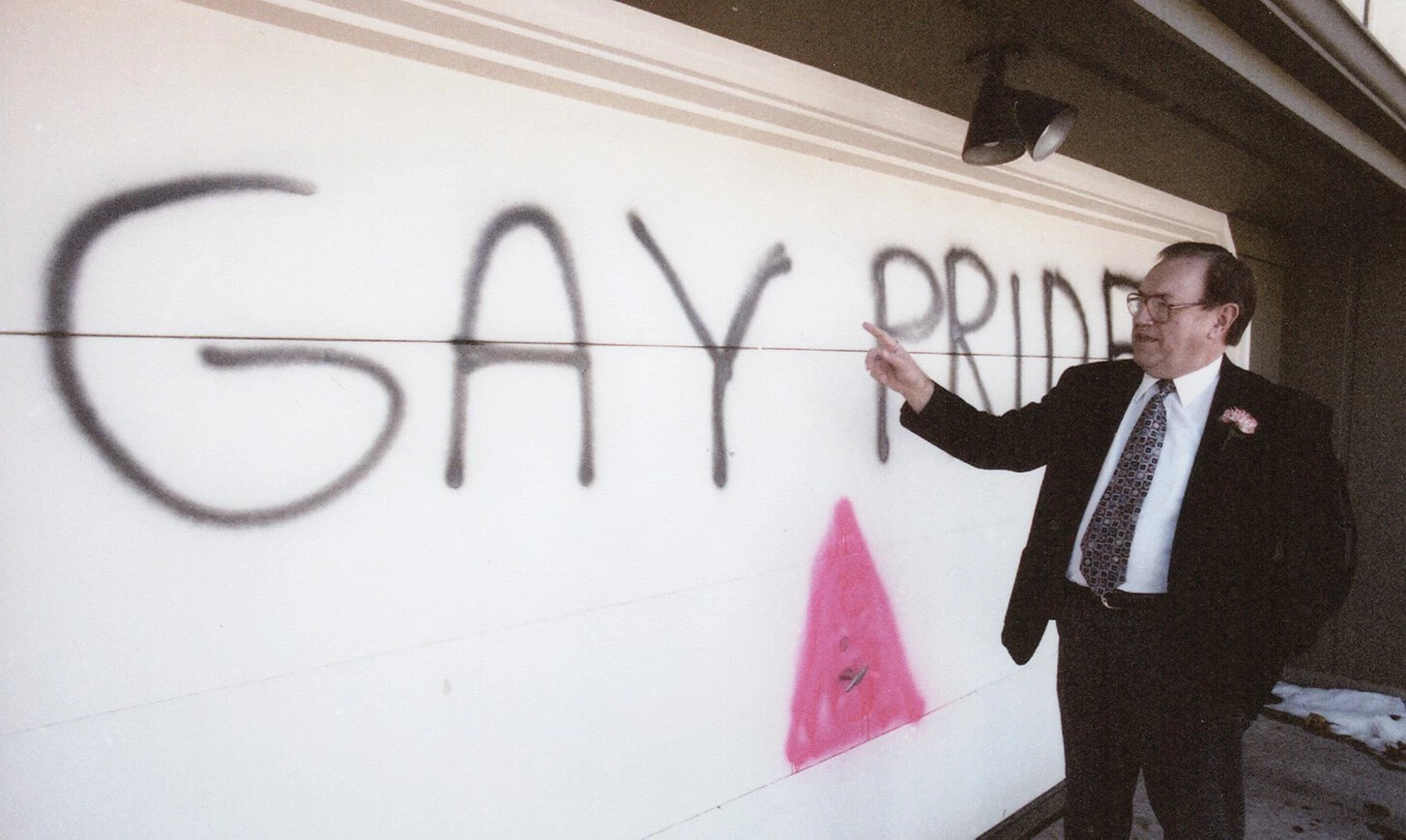 White man in a suit and tie pointing at spray paint on a white garage door.