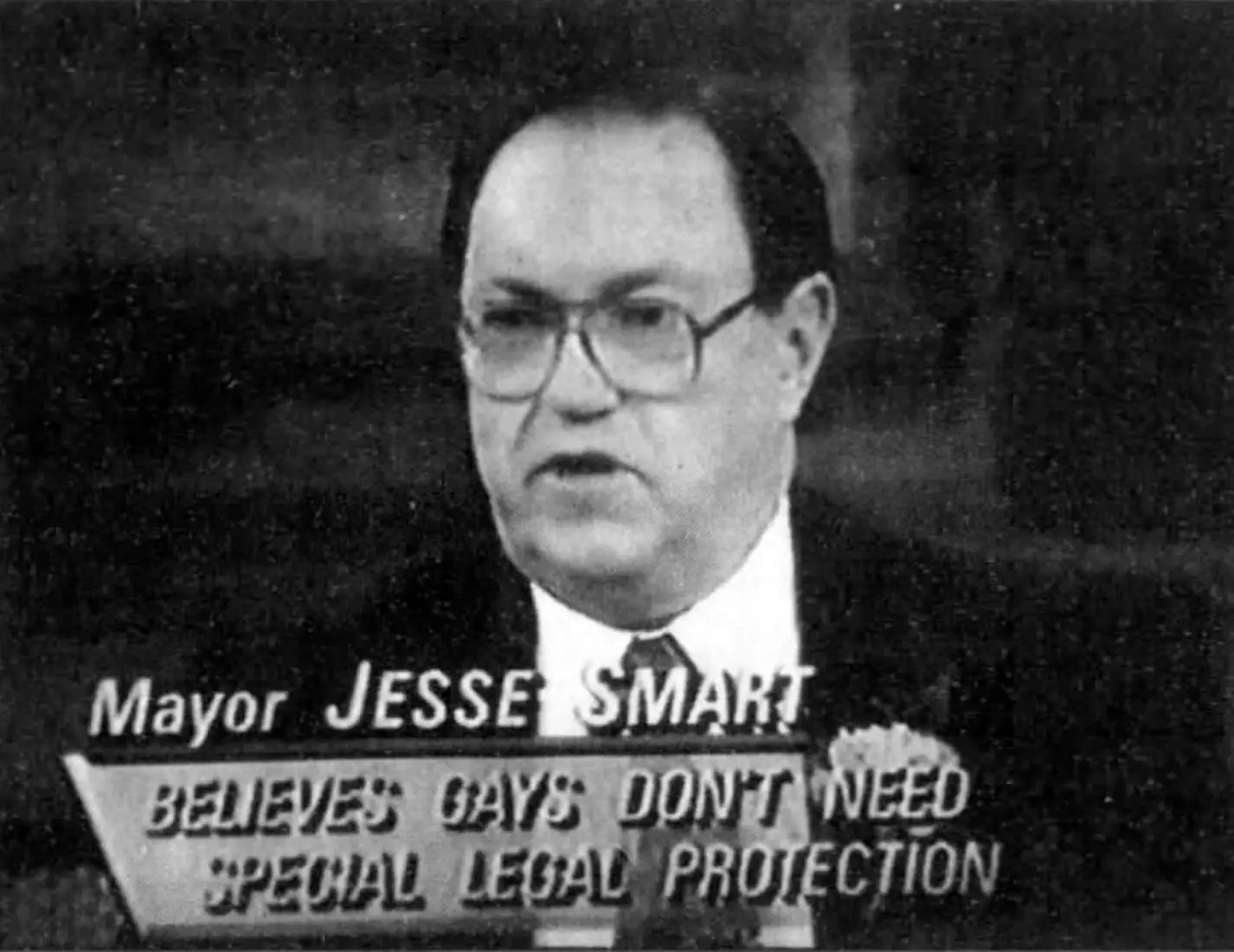 A black and white still shot from a TV screen showing an older white man wearing a suit, tie, and glasses.