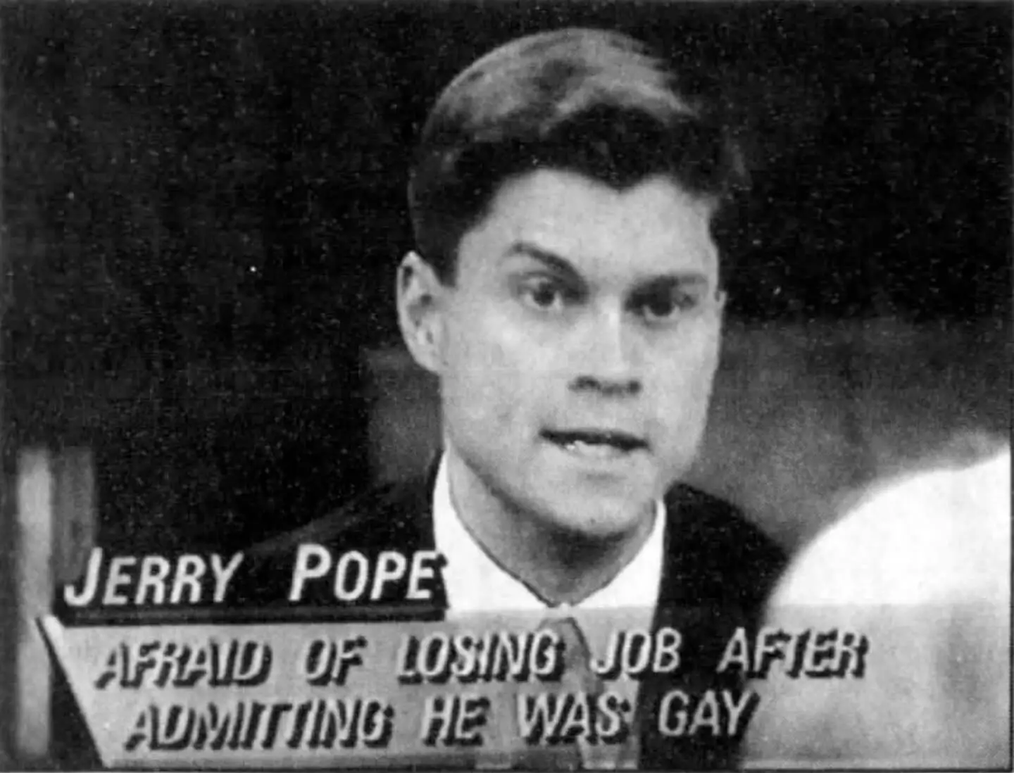 A black and white still shot of a TV show showing Jerry Pope, a white man with dark hair wearing a suit and tie.