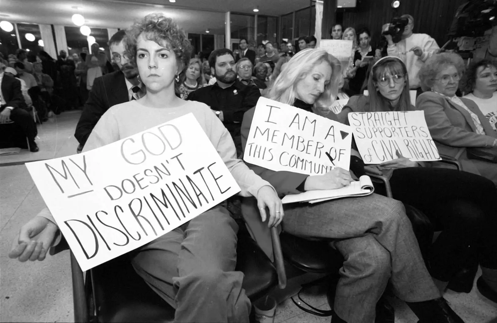 A seated crowd at a public meeting, Three women in the front row are holding poster board signs.