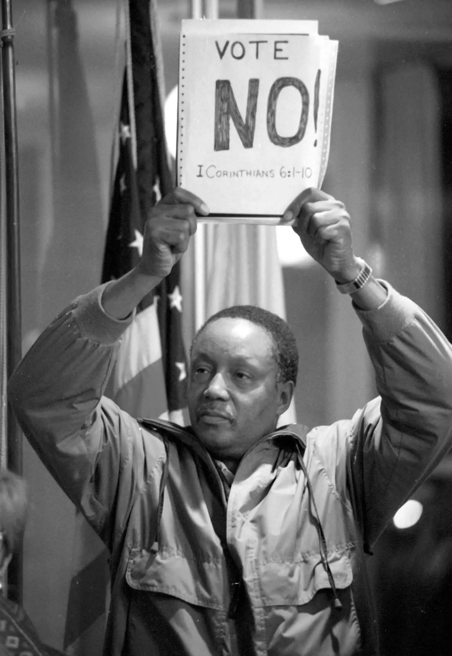Black and white photo of a Black man wearing a jacket holding a sign over his head.
