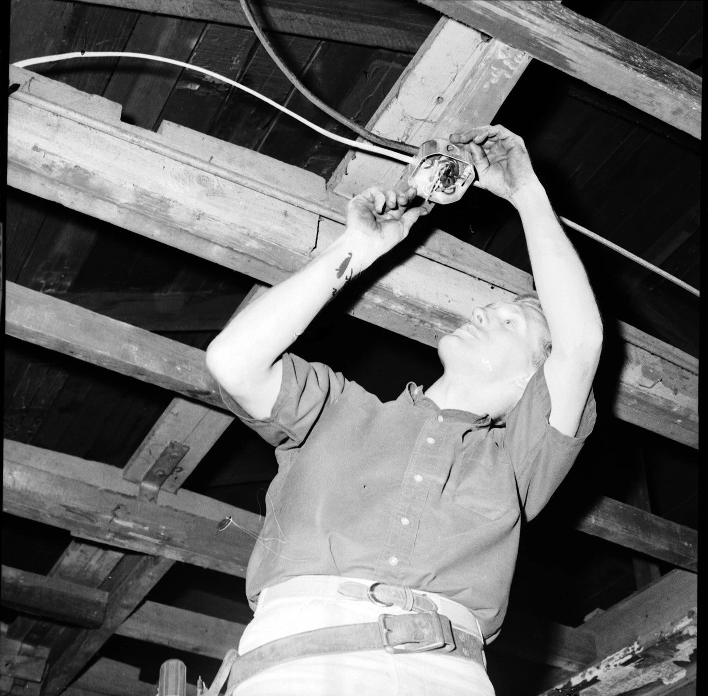 Black and white photograph of a man doing electrical work on a the ceiling rafters of a home.