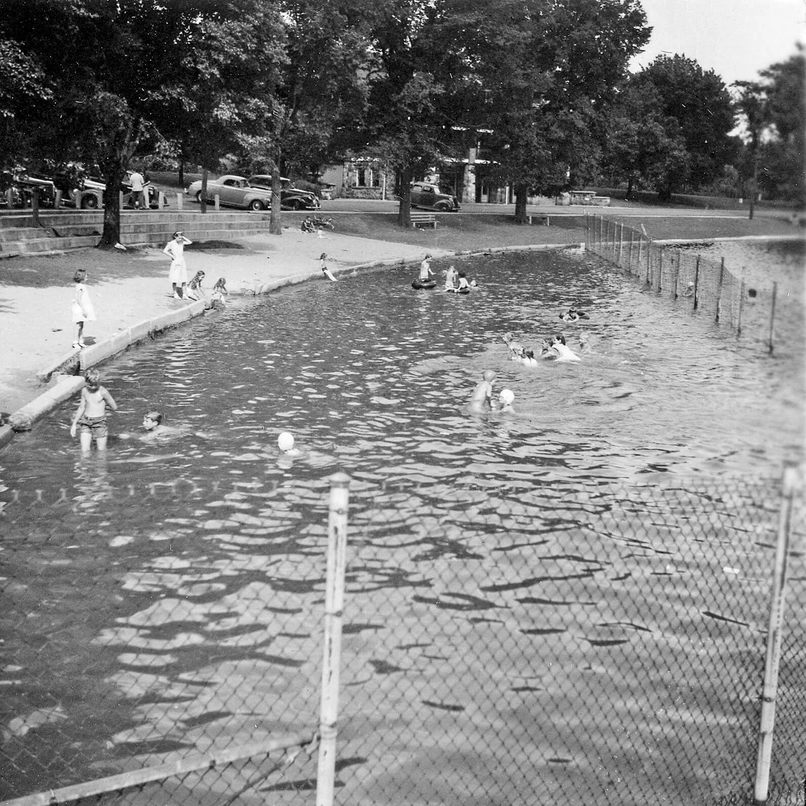 Black and white photo of a lake shoreline with sandy beach, full of children swimming with their parents. A chain link fence divides the deep area from the shallow area.