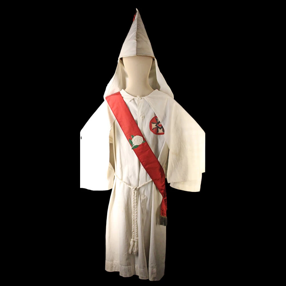 Mannequin wearing KKK robe. There is a wide red ribbon going across the chest. There is a white rose on the ribbon. There is a white rope tied at the waist. There is a hood that comes to a point at the top.