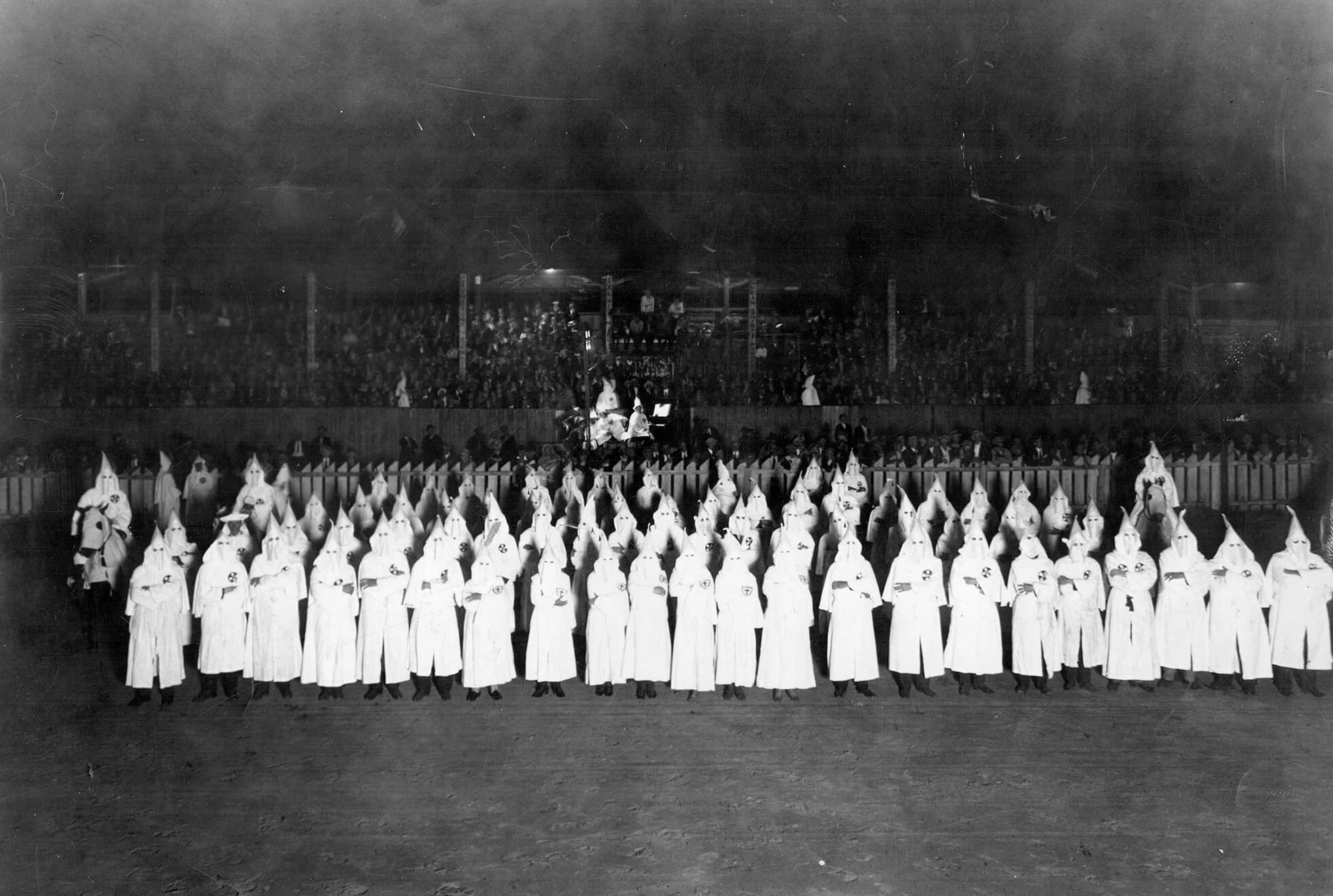 Black and white photo of large group of robed klansmen, lined up in rows. There are approximately 75 people in robes and hoods, and some men are on horses. In the background there is seating, like a stadium, and they appear to be full of people.