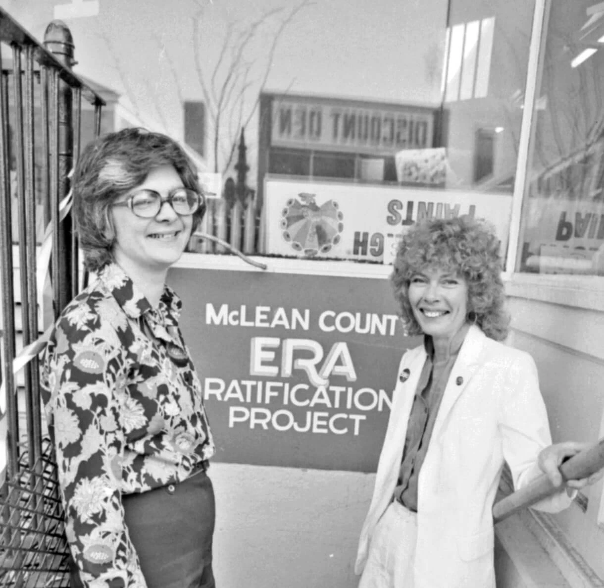 Two white women are standing at the top of a staircase with a sign behind them supporting the ERA amendment.
