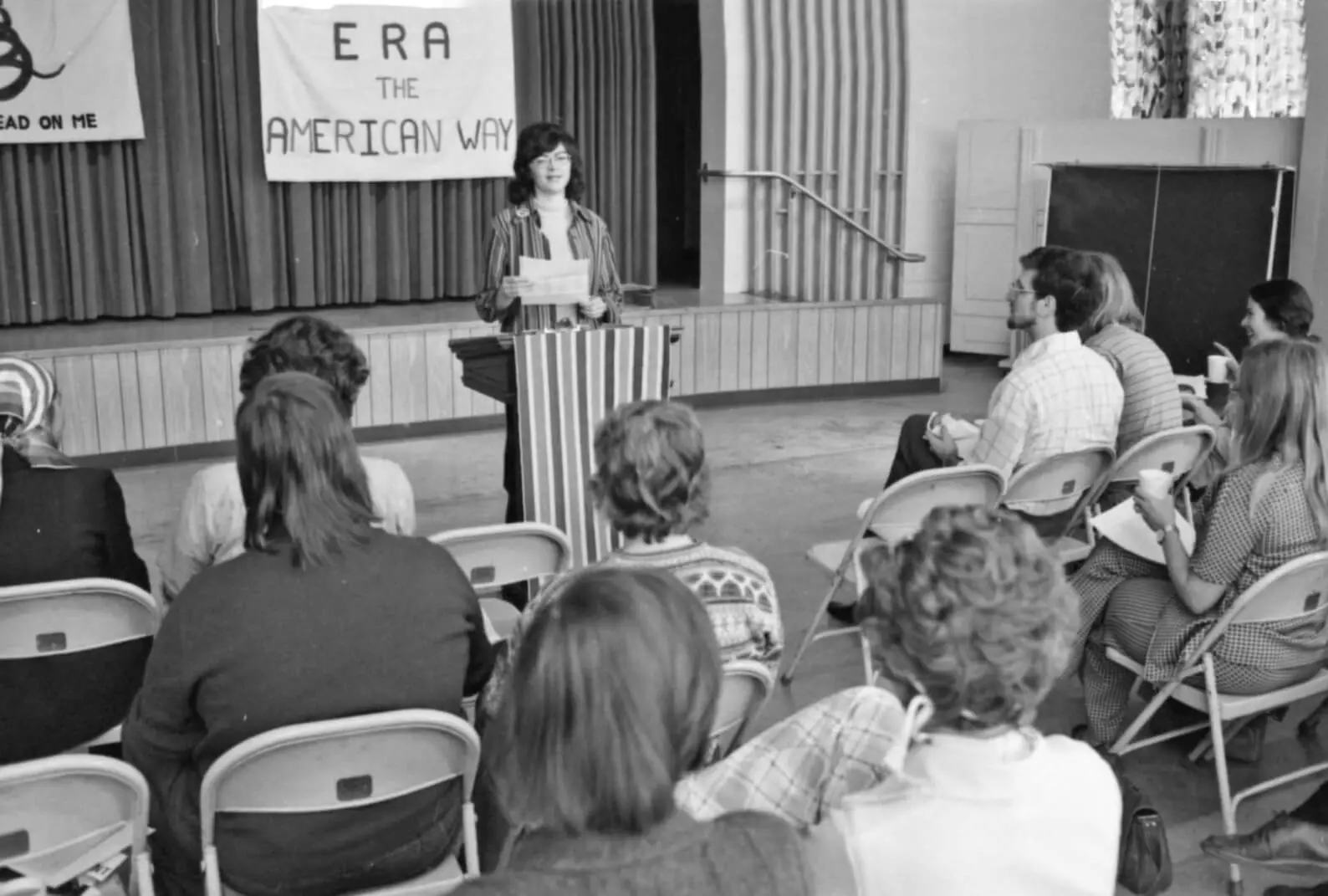 Black and white photo of a white woman standing at a podium in front of a small audience.