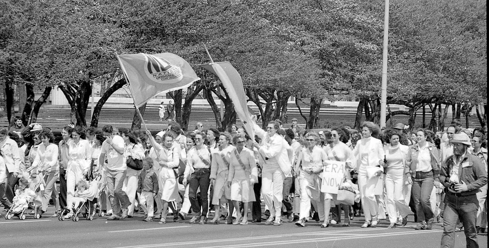Black and white photo of dozens of women (and a couple men) wearing white are walking in a crowd carrying flags. They are wearing sashes and smiling. Some are pushing strollers.