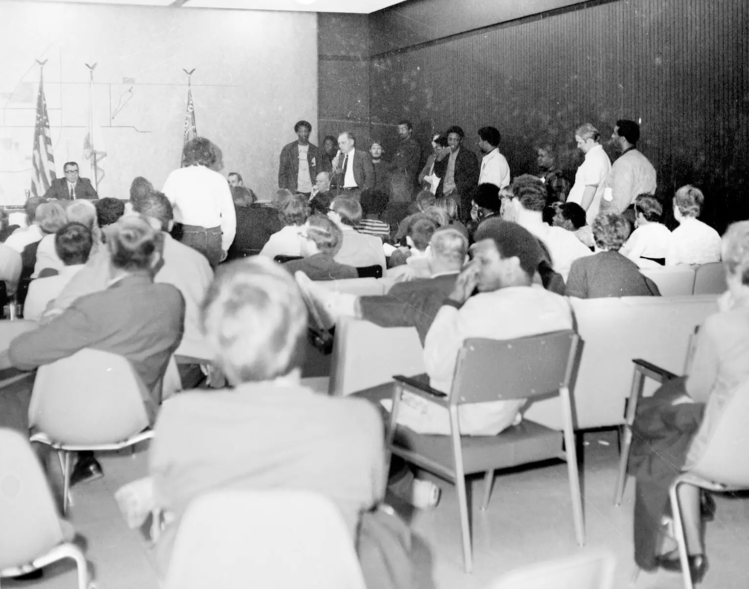 People are seen sitting and standing listening to a speaker at a city meeting.