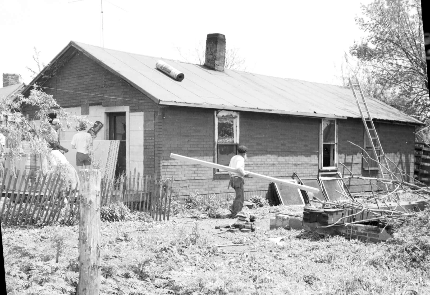 There is a small one-story home.  A man is carrying a board toward a ladder which is leaning on the roof.  A roll of roofing is on the shingle-less roof.