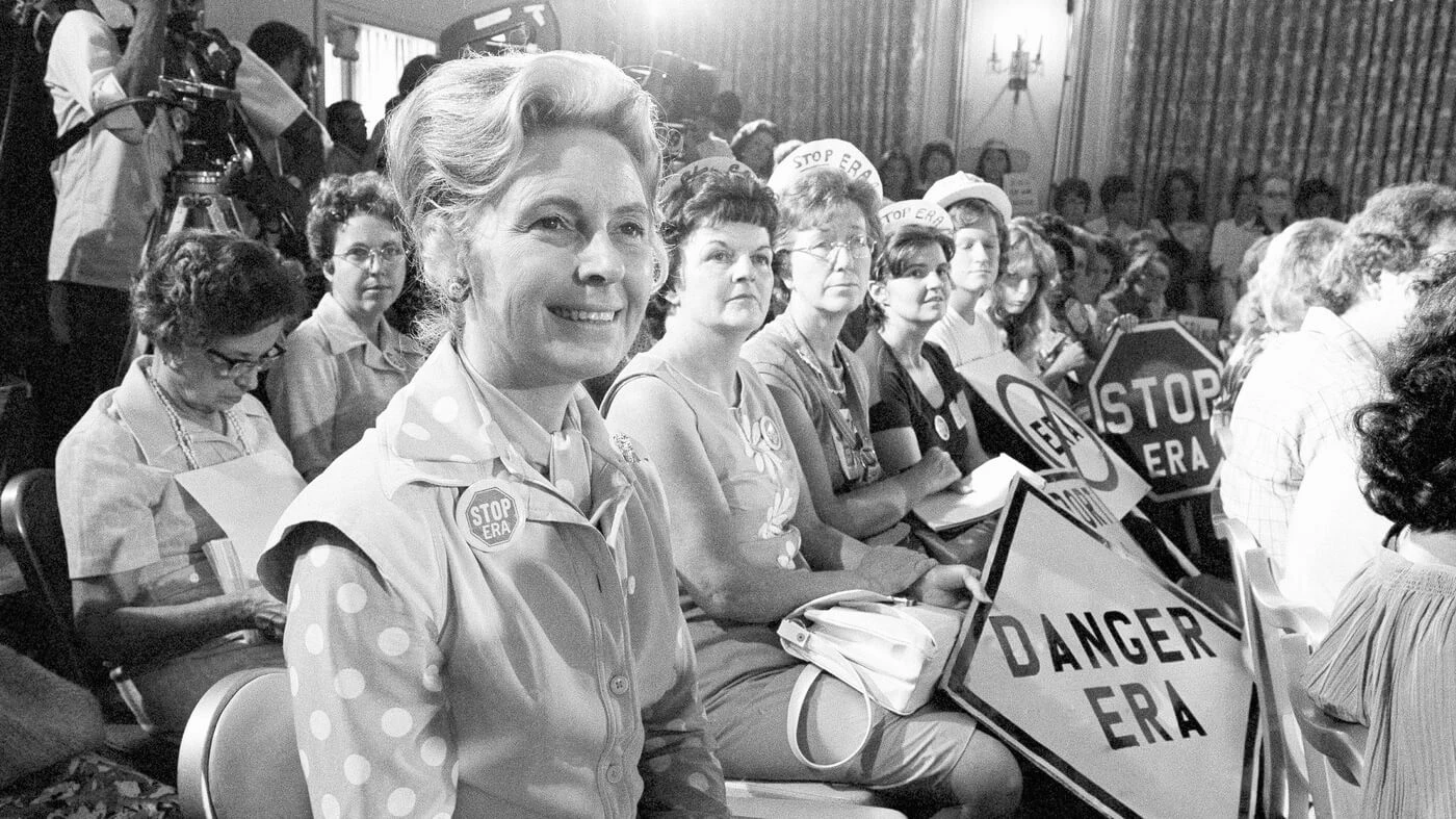 Black and white photo of a group of white women seated, holding signs against the ERA.