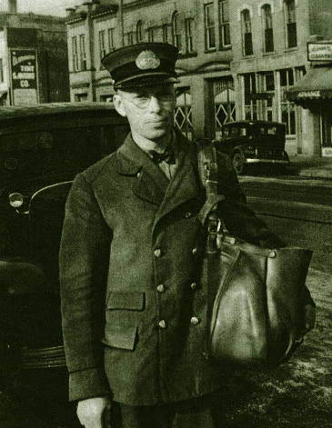 A light-skinned man in a buttoned up jacket, bow tie, and mail carrier cap stands outside near a street. He is carrying a large leather bag on his left shoulder.