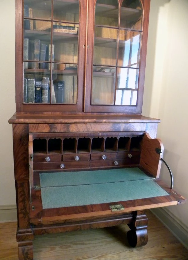 American Empire style secretary desk with gothic arched glass doors on shelved bookcase at top. Doors have brass eschutchins and moulded trim around top section. Inside the doors are three shelves. A  hinged panel opens to provide writing surface in front of pigeonholes and little drawers with glass pulls. Below the writing surface are two 2 drawers. At the bottom of the desk are four large scroll or rounded feet.