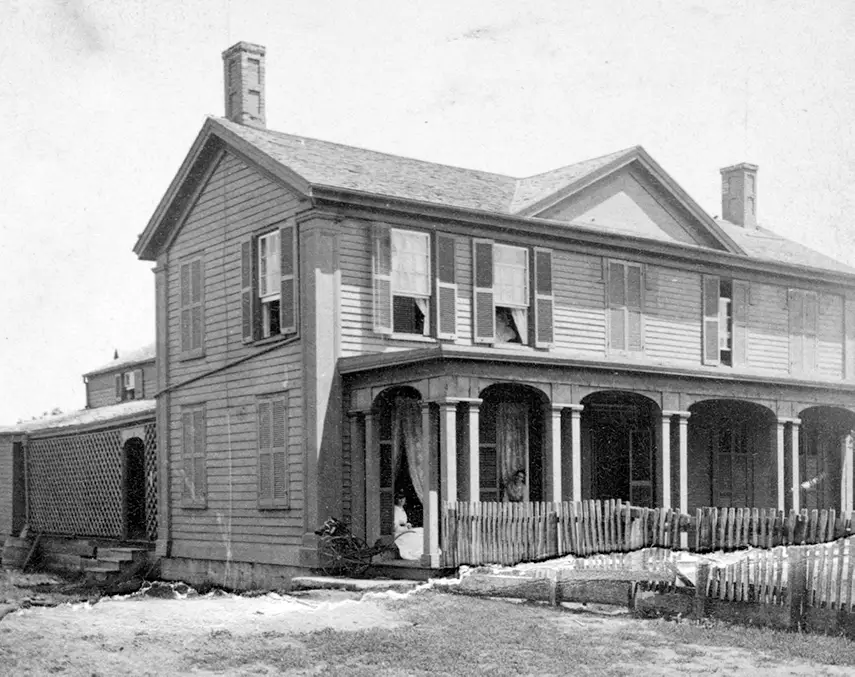 Black and white photograph of the Davis home, two stories in size, with a wide front porch and several windows with open shutters. A woman sits on the porch in the window frame and a man stands close by, slightly hidden by a fence in front of the columned porch.