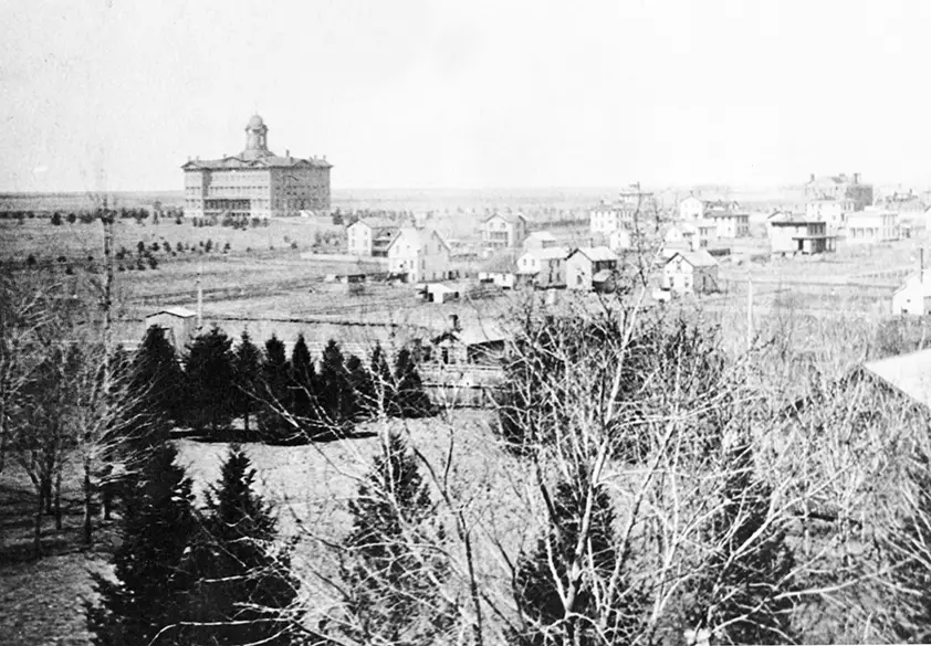 Black and white photograph of Normal. Trees are closest in the frame, separating the house from the other houses in the town. In the upper left corner is a bigger building with a bell tower.
