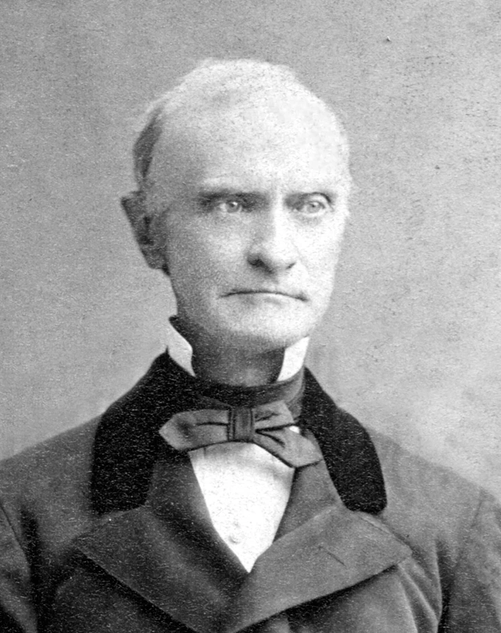 Black and white photo of an older man, with suit and tie and white collar. He looks away from the camera with a downturned smile.