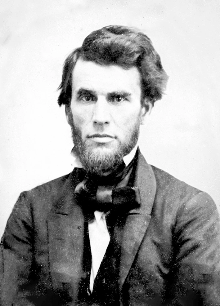 Black and white photograph of a young man in a suit. He has thick hair and a full beard, his eyes look directly into the camera.